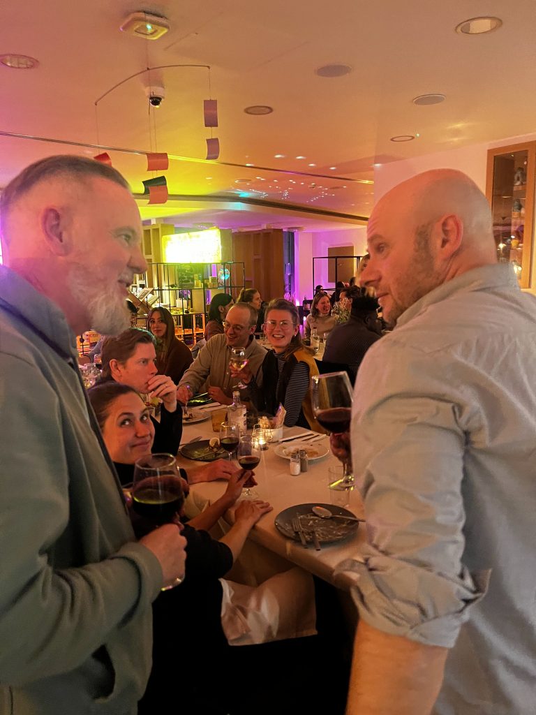 A bunch of people chatting in a bar location, two men in the foreground on either side chatting to each other