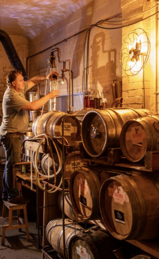 A man in a dark distillery with barrels of beer is fixing some pipes