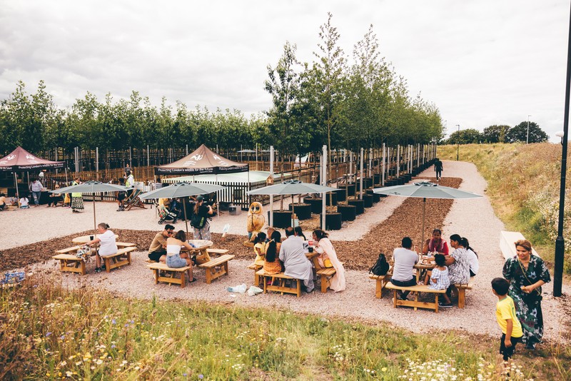 Several people sit around four wooden picnic tables with umbrellas, in front of them and to the right is a green grassy and flowery bank, behind them are green trees and more large umbrellas