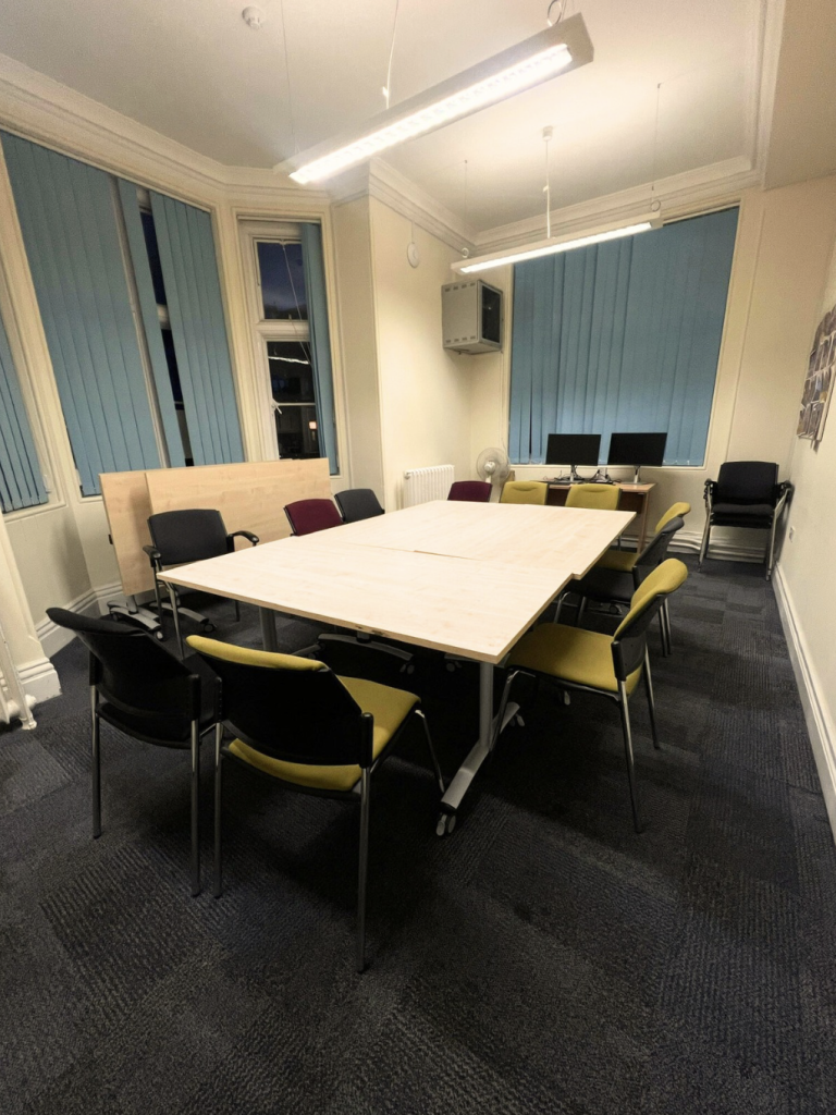 Ten chairs around a conference table in an empty room with cream walls, blue shutters on the window and grey carpet