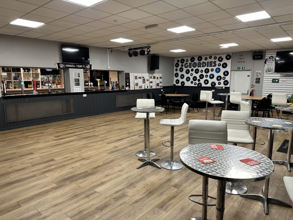 Indoor space with bar along one end. A few circular tables with bar stools. Wooden floor and square lighting in false ceiling.
