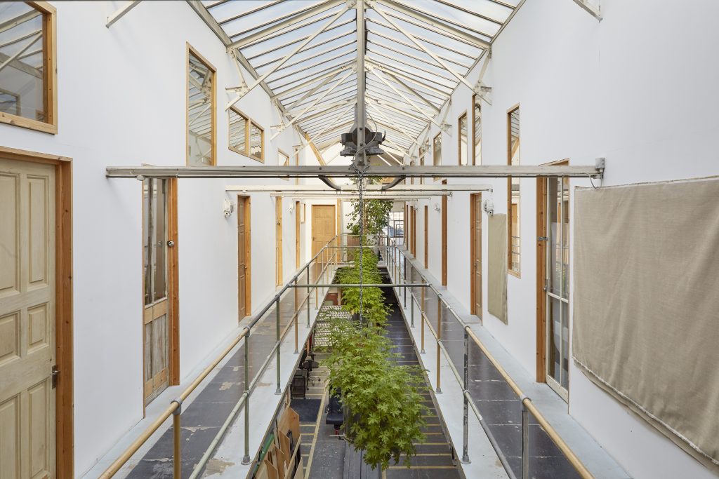 A long, airy interior space with a glass apex ceiling, greenery below, and steel walkways either side of an atrium with wooden doors set in white walls.