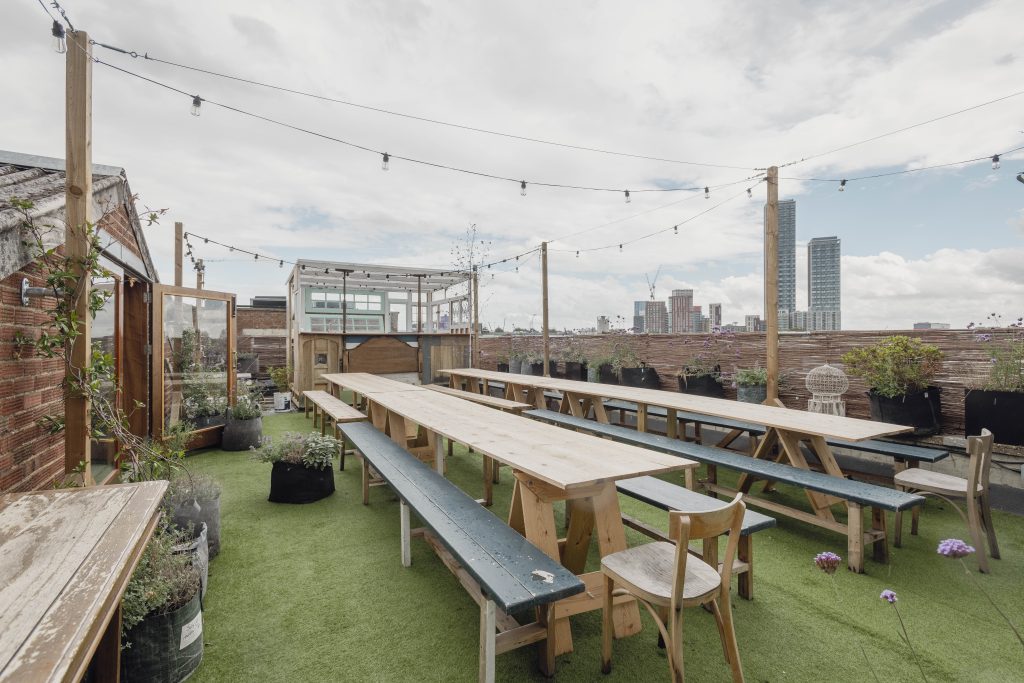 A bar area on a roof with green floor covering and long benches. Some lights strung overhear and clouds in a blue sky.