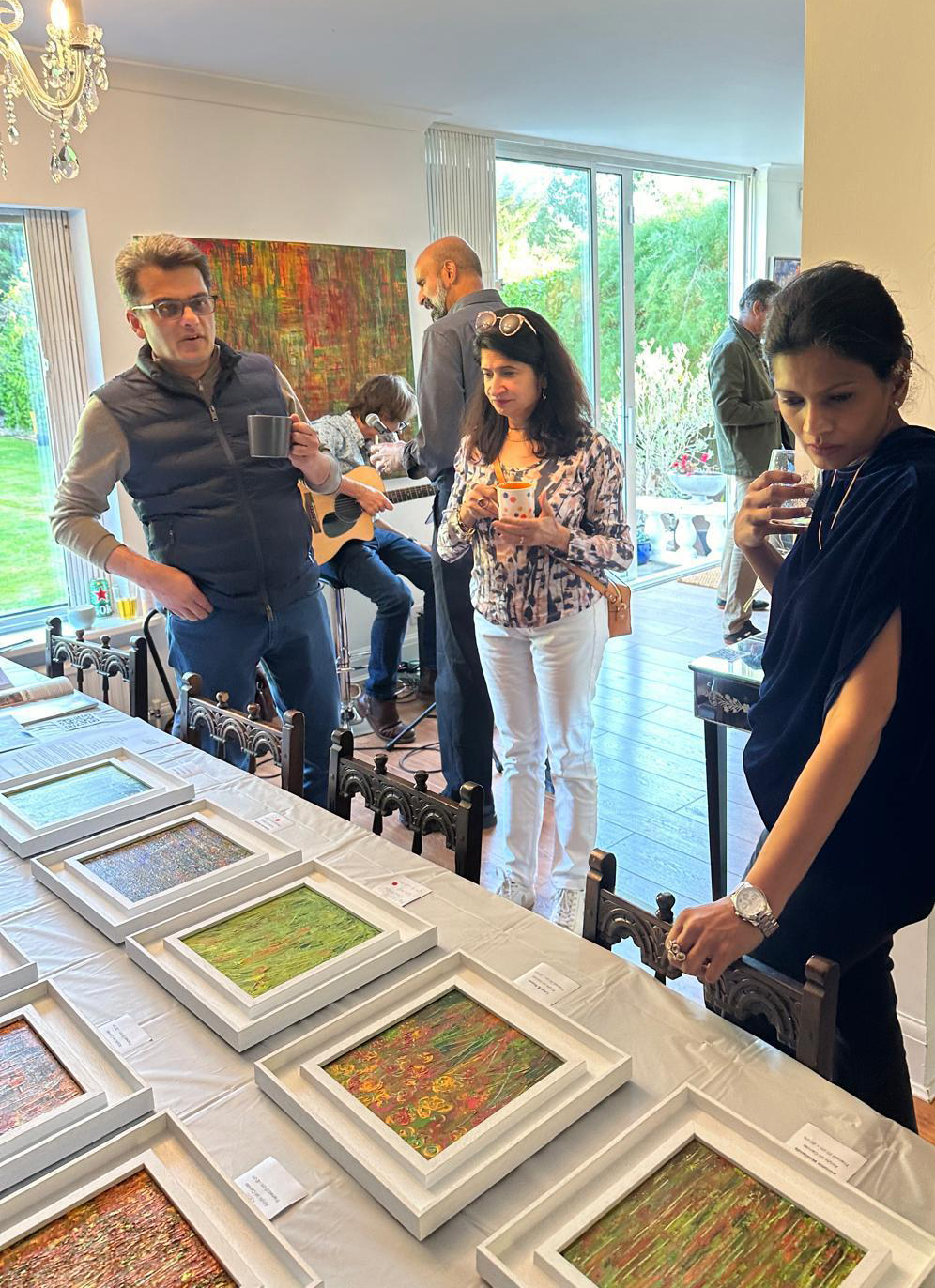 A few people in a room well lit with sunlight look at a number of framed images on a table as someone in the background plays guitar