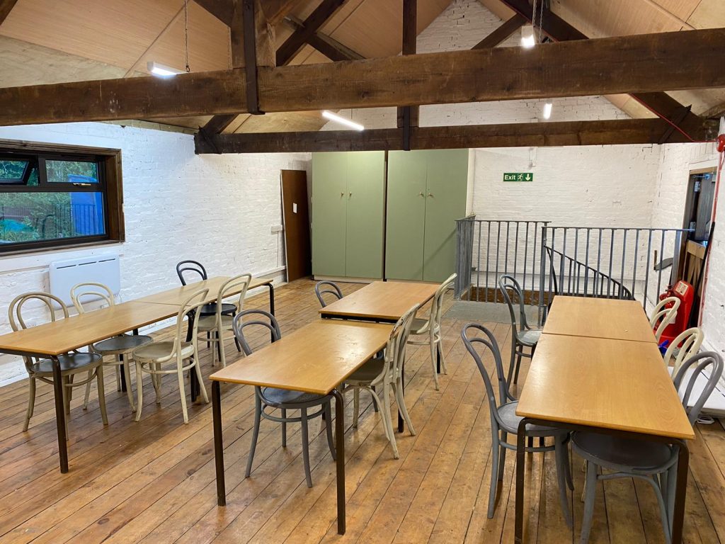 Indoor space, wooden floors, tables and chairs arranged in rows, classroom style. Rafters overhead.