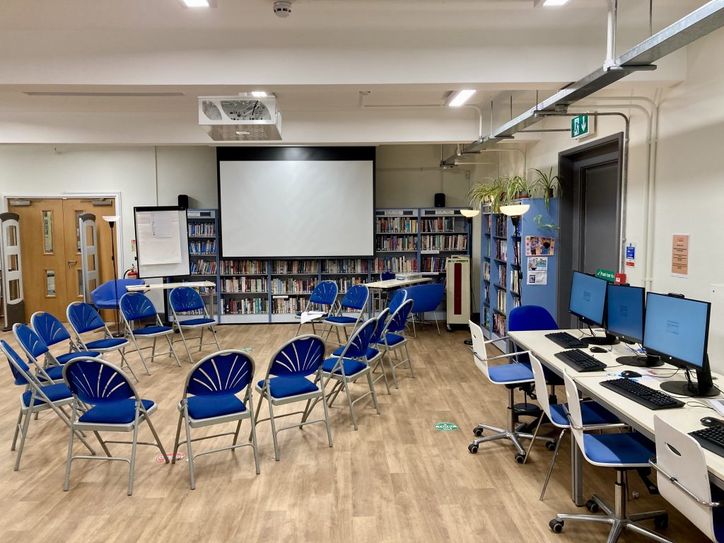 A room with white walls and ceiling and a wooden floor. Blue chairs arranged in a circular formation and a screen at one end.