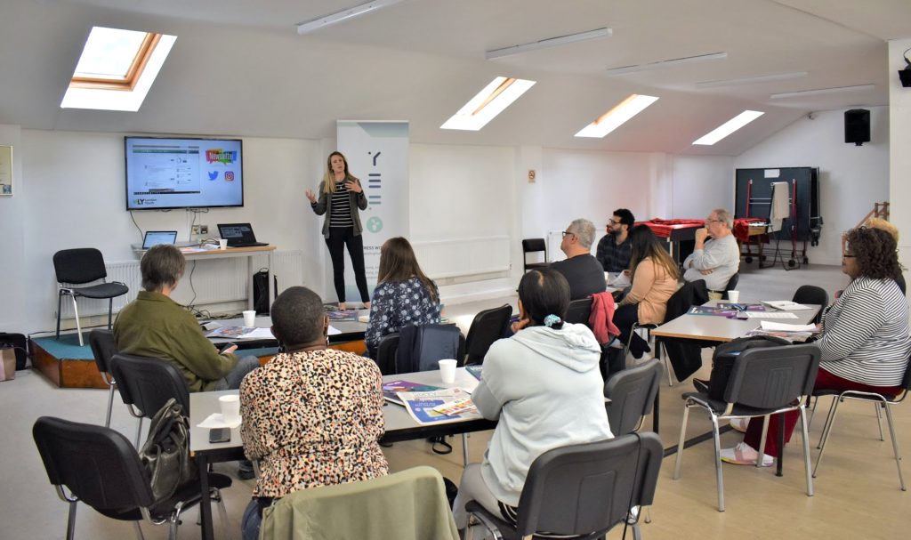 Indoor learning space, people at tables facing a woman standing up next to a screen.