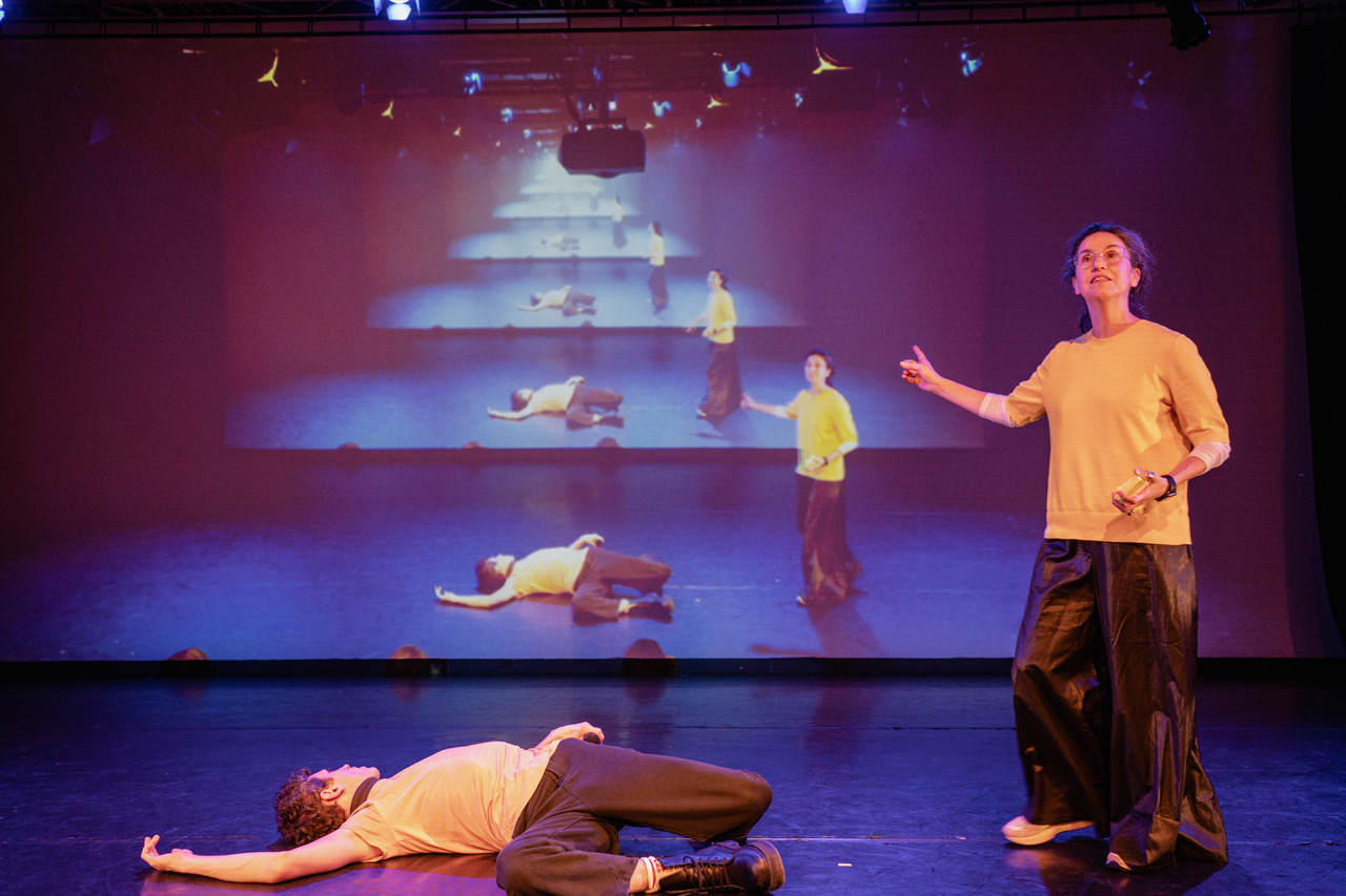 A stage with two performers wearing orange tops and black trousers, behind them is a screen with images of them recurring into the background
