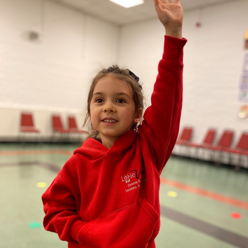 A young girl in a red hoody stands with one hand in her pocket and the other raised in the air