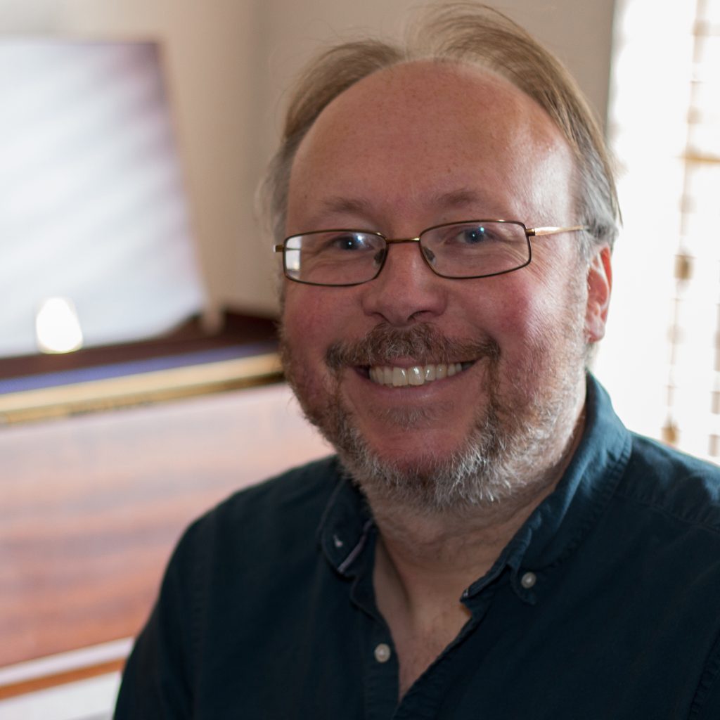 A man with glasses smiles at the camera against a blurry background that is cream and may have a brown piano