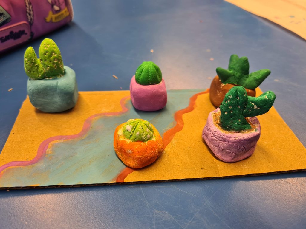 Five clay sculptures of cacti in pots sit on a colourful painted piece of slate