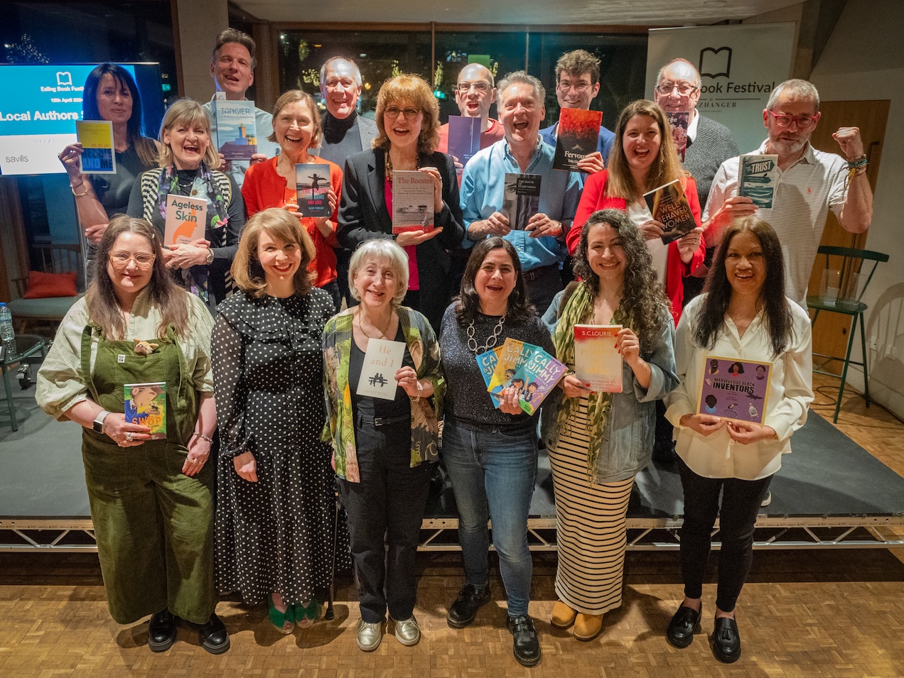 18 people posing for the camera standing in three rows. They are all smiling and holding different books.