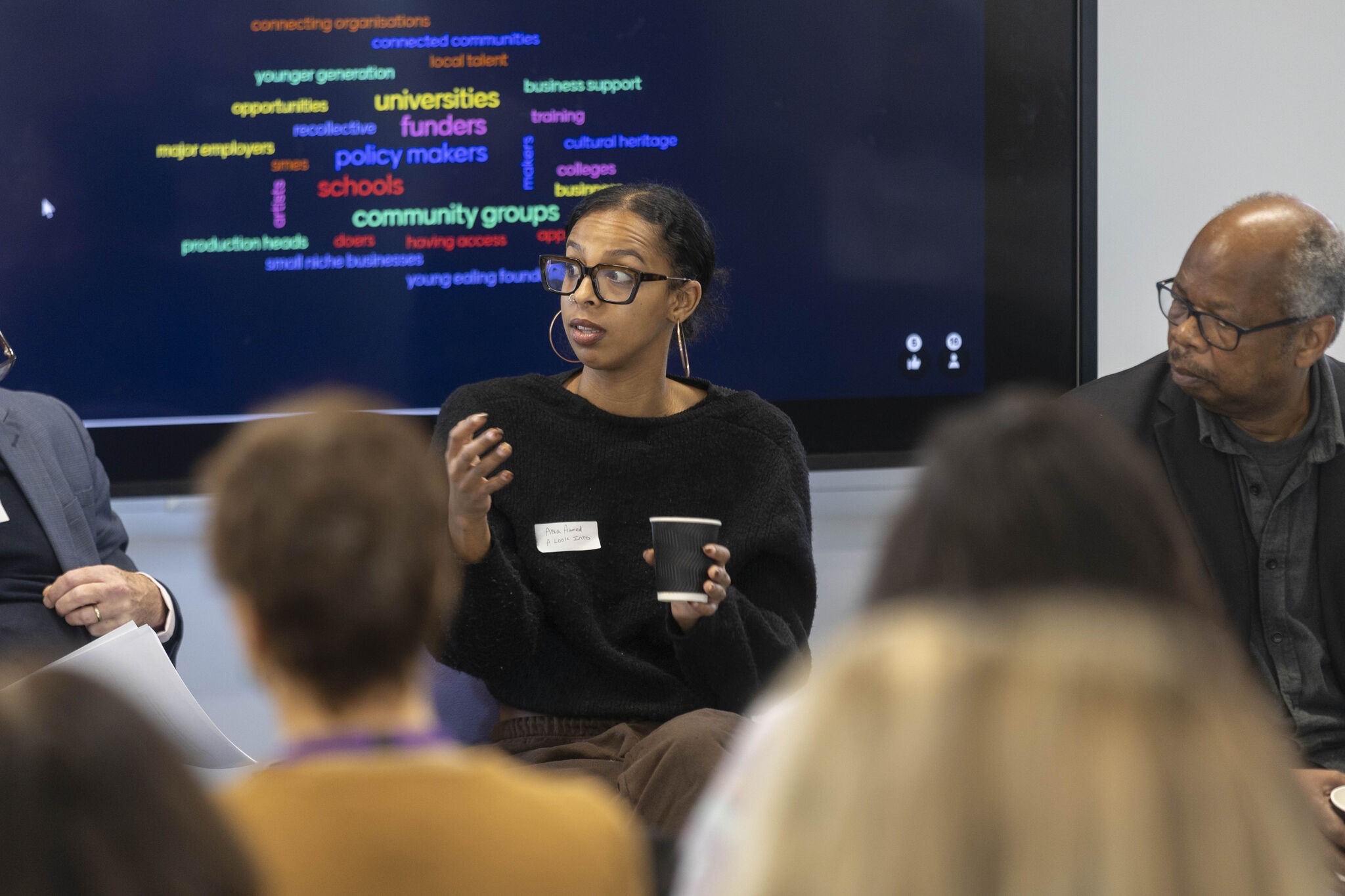 A woman with black glasses is sat speaking in front of a screen with lots of colourful words on. To the right a man sat next to her looks on and in the foreground there is an audience.