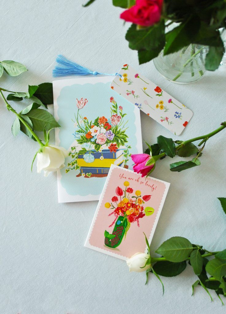 Looking down on two greetings cards, a book mark, a vase with roses in and three roses strewn across the white table