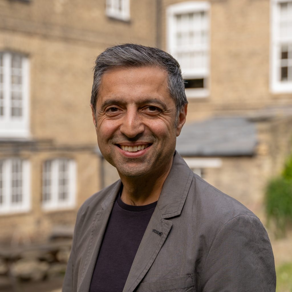 a man smiles and looks at the camera, he wears a suit and is stood in front of a limescale building