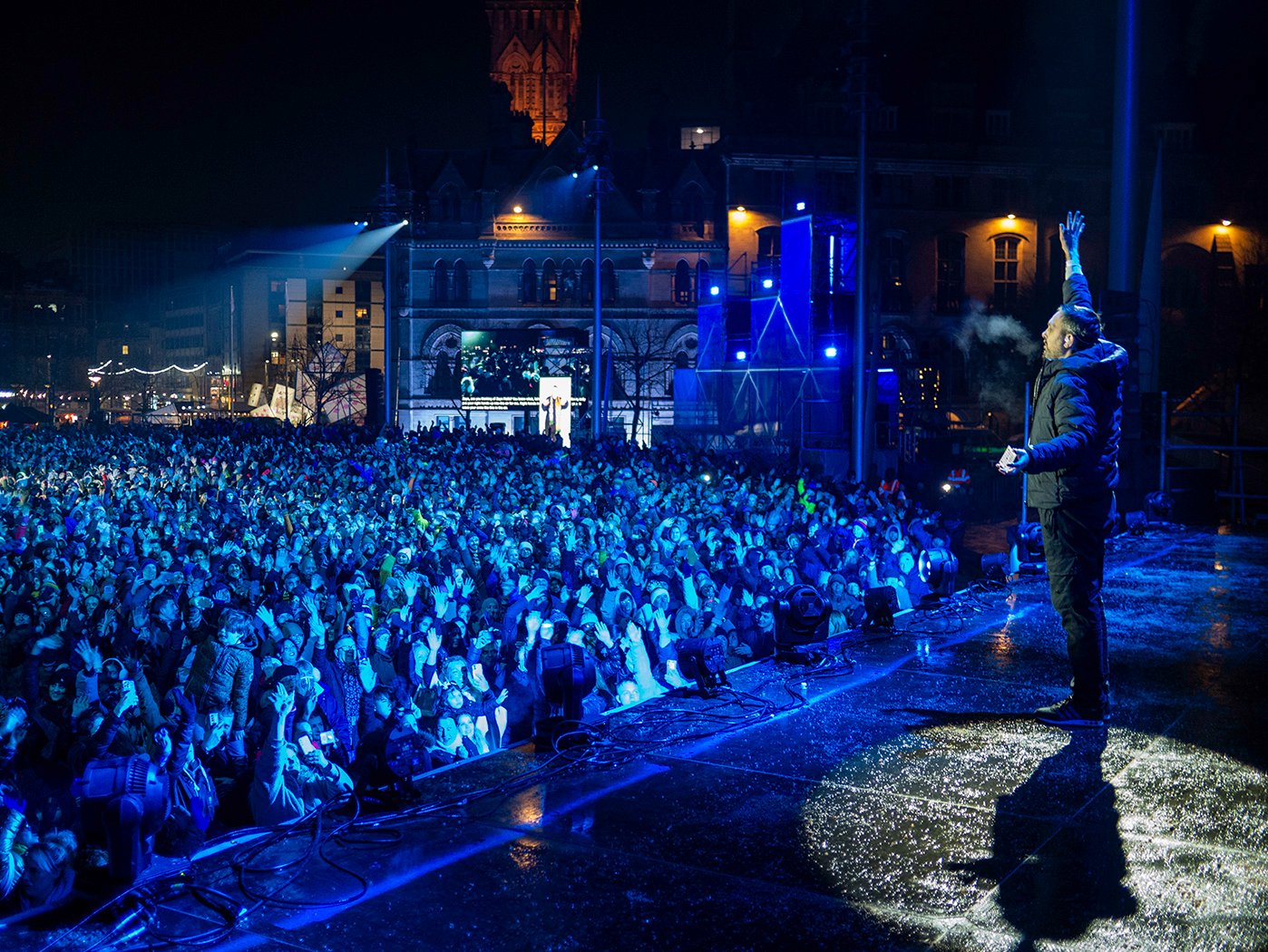 A person stands on stage with his hand up in front of a packed audience covered in a blue light
