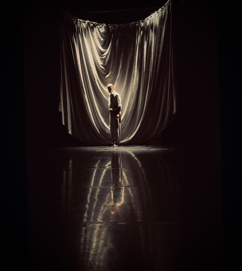 A dark room, someone stands in the distance, dimly lit and surrounded by large curtains