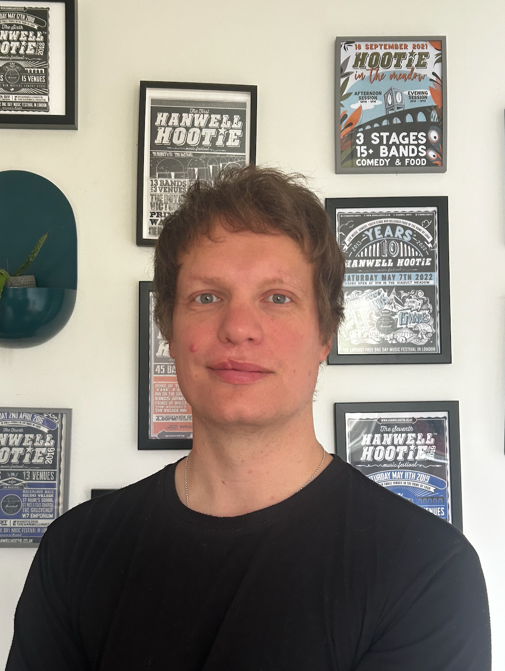 A man with fair hair smiles gently and looks into the camera. He is stood in from of a white wall with lots of black and white pictures hanging up.