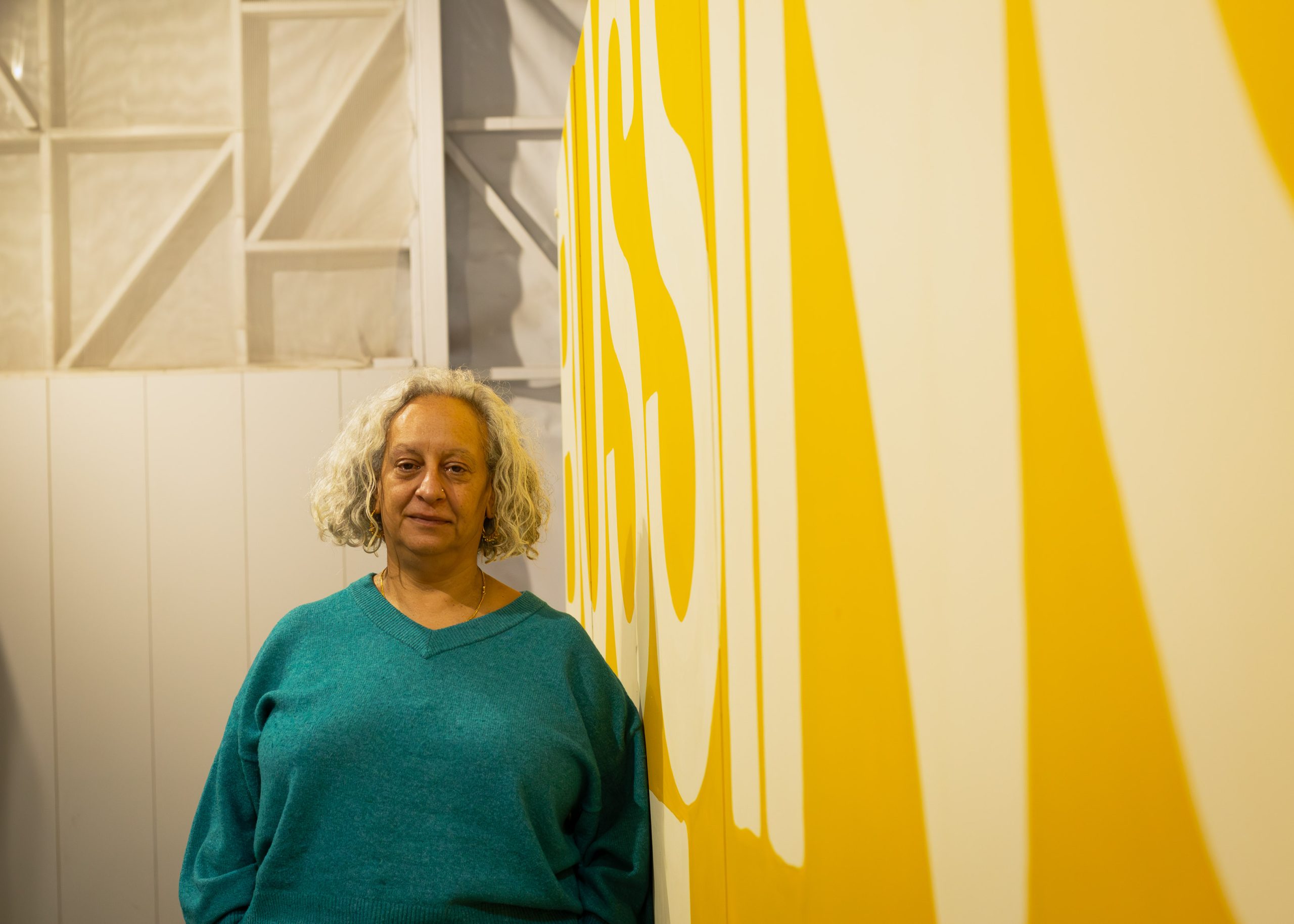 A woman with bobbed blonde hair leans onto a wall with a dark and light yellow design with some illegible letters.