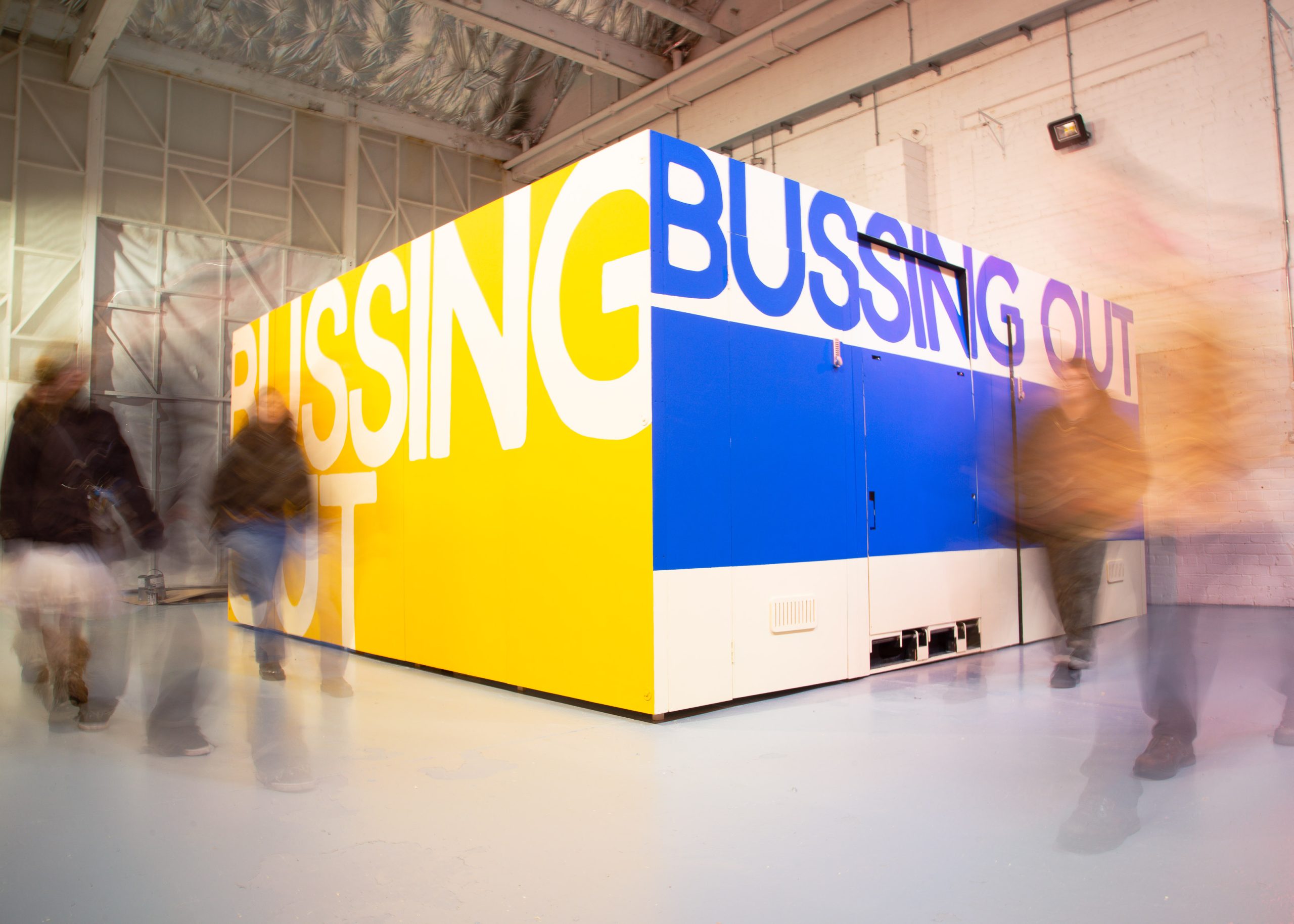 A number of blurred moving people in a large white studio with a huge cubeb in the middle with yellow and blue designs with the words 'Bussing Out'