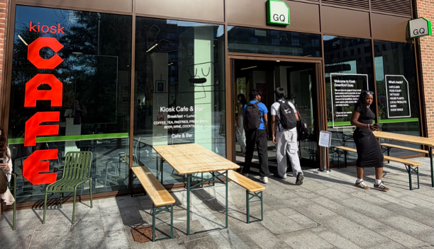 The outside of a cafe on a sunny day. A few people mill about outside and red letters read CAFE on the window on the left.