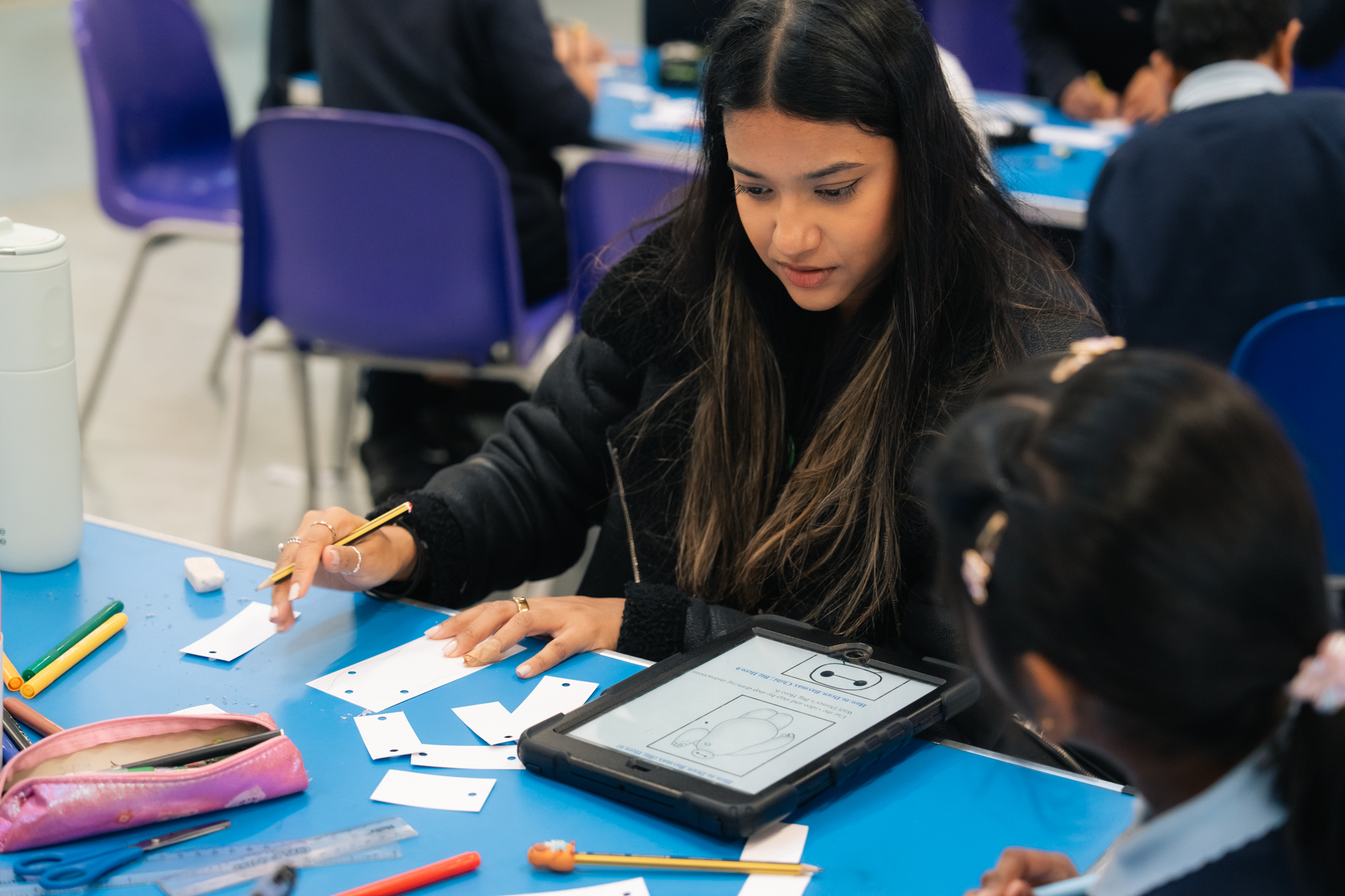 A room of students sat at blue tables on blue chairs. In the foreground two girls are making arts and crafts