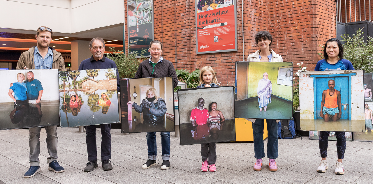 Six people stand in a line next to each other. Each is holding a large print of a photograph. They are stood in a shopping centre.