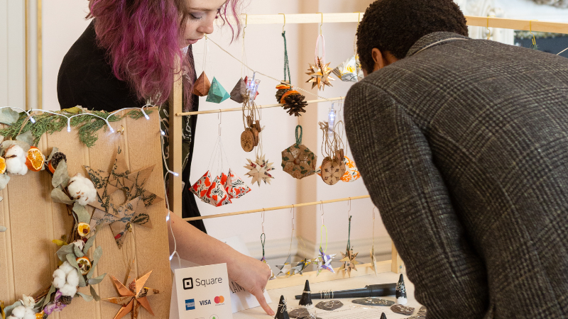 A craft stall with a maker behind a table and a man looking on at her colourful Christmas trinkets