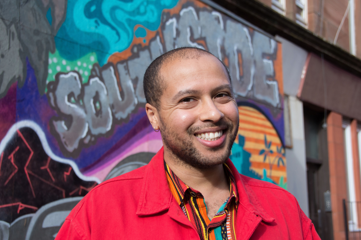 A man in a red shirt smiling at the camera. He stands in front of a wall with lots of graffiti on.